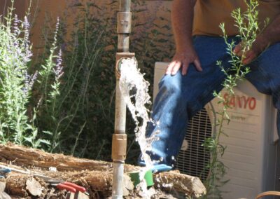 Water bursts from a valve on a pipe coming up from the ground. Behind it a worker sits next to a stucco wall observing.