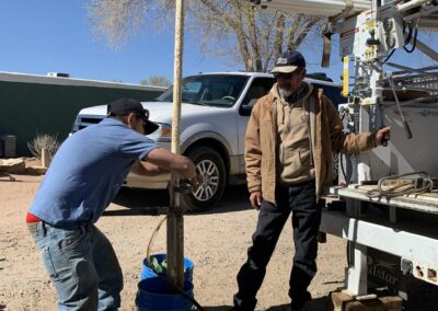 Two workers stand at the rear of a crane truck. One observes while the other is working on the pipes attached to the crane. A large pipe from a well rises from the ground in front of the two men.
