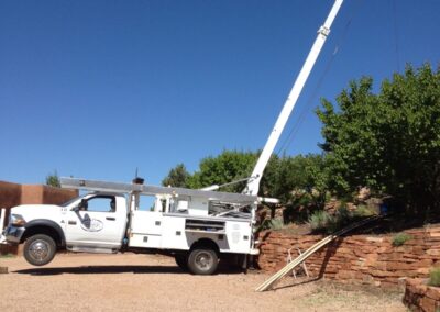 A crane truck is backed up to a stone wall. The crane is lifting something that is hidden by a tree in the foreground. The front wheels of the truck have lifted slightly off the ground.