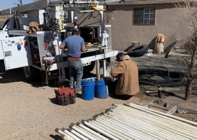 A crane truck is parked and the rear tool area is open. One worker is looking at the tools, while another sits on the ground working on something in his lap. A large number of pipes are stacked in the foreground.