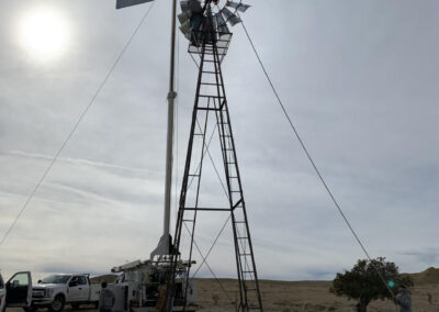 Workers stand around a windmill. On e holds a rope attached to the top of the windmill. A crane truck sits behind the windmill.